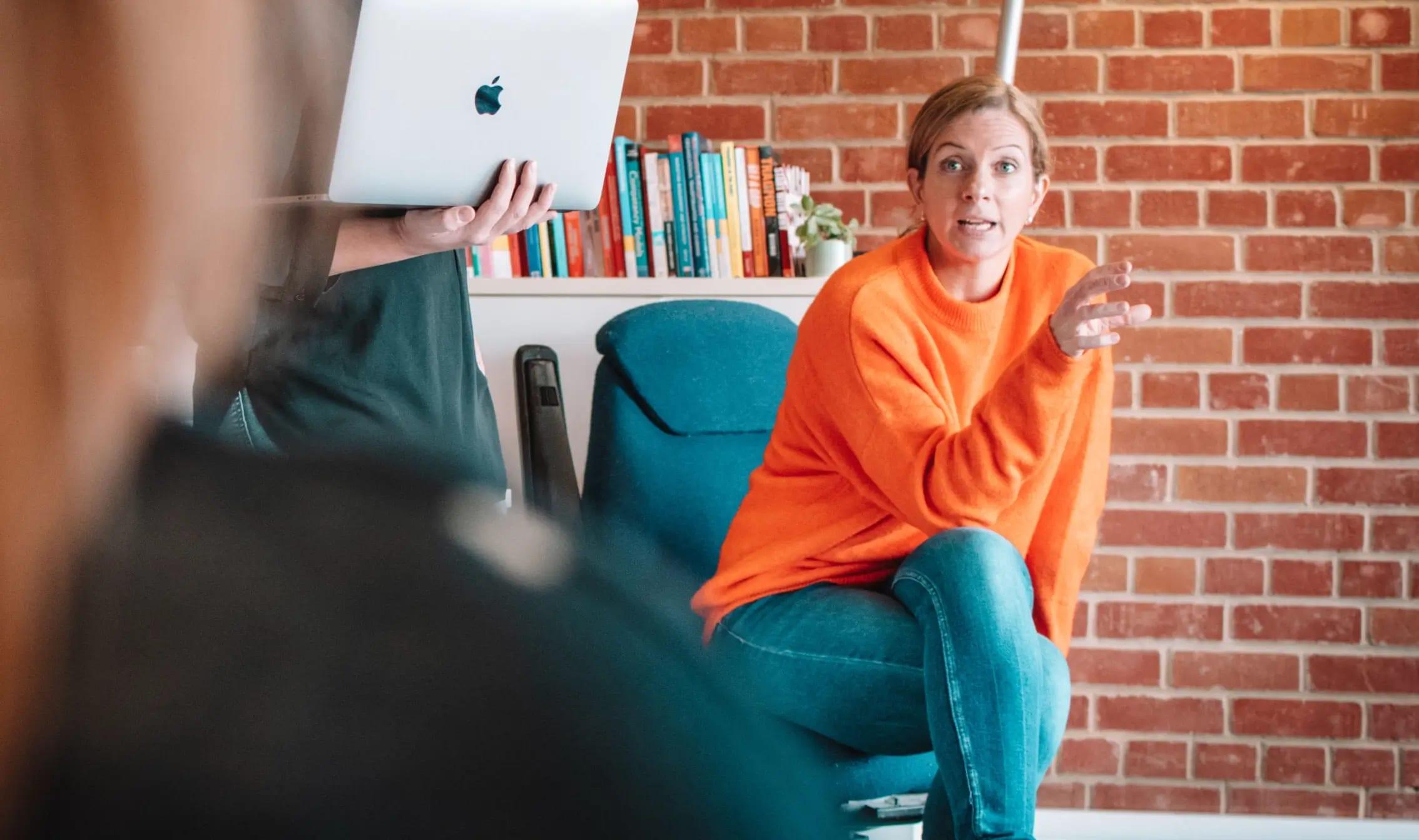 person sat on an office chair directly addressing a room of people and gesturing with their hand 