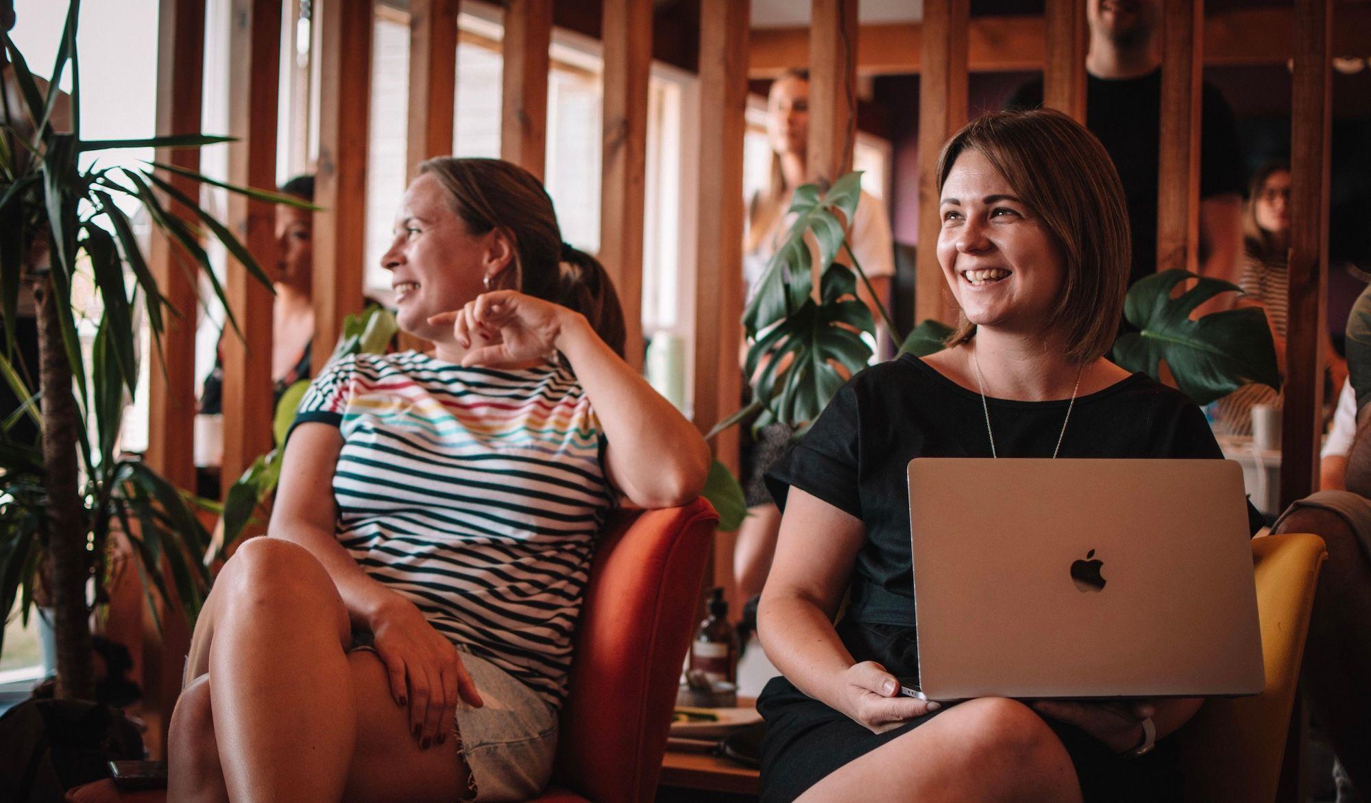 Two people sat on chairs laughing. One of them is holding a laptop in their hands.