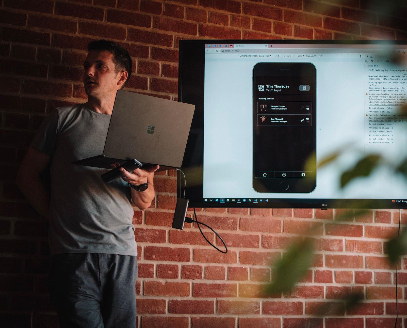 Person stood presenting in front of a tv screen with a laptop in their hands.